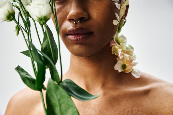 Ένα κοντινό πορτραίτο ενός νεαρού μαύρου με λευκά λουλούδια που στολίζει το πρόσωπο και το σώμα του πάνω σε γκρι φόντο.. - Φωτογραφία, εικόνα