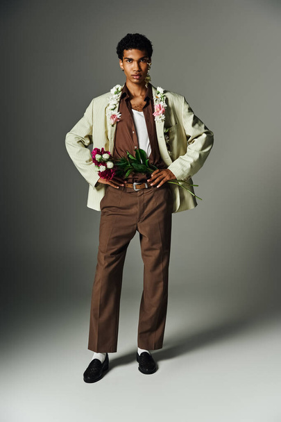 A young African American man in a cream blazer and brown pants poses with flowers on his neck and face against a grey background. - Fotografie, Obrázek