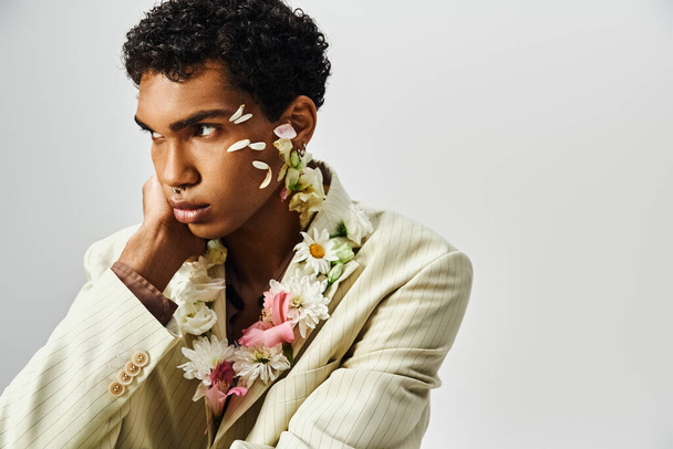 Un giovane uomo afroamericano con fiori che adornano il viso e il collo posa in un blazer elegante su uno sfondo grigio. - Foto, immagini