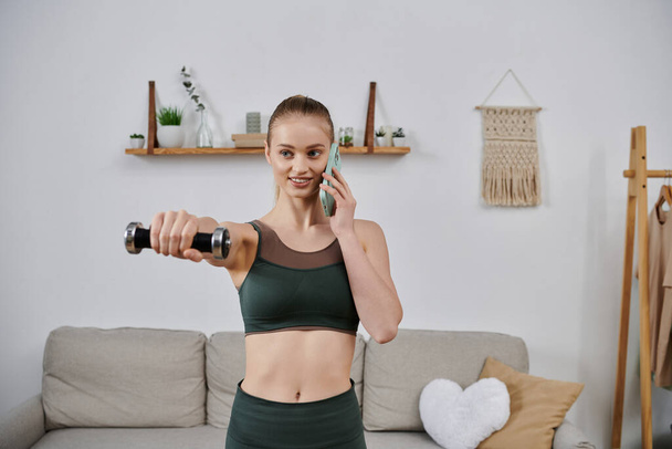 Молодая женщина в спортивной одежде упражнения с гантели, разговаривая по телефону дома. - Фото, изображение