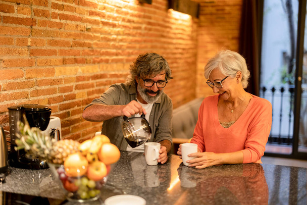 Mutlu son sınıf çifti kahve yapıyor ve tuğla duvarlı bir mutfakta gülümsüyor.. - Fotoğraf, Görsel