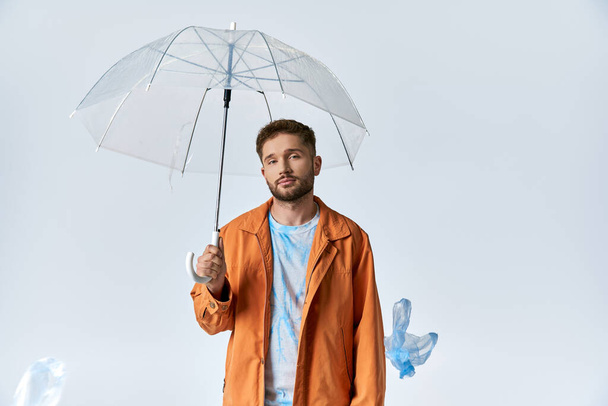 A man in an orange jacket stands under a clear umbrella, with plastic objects surrounding him. - Fotó, kép