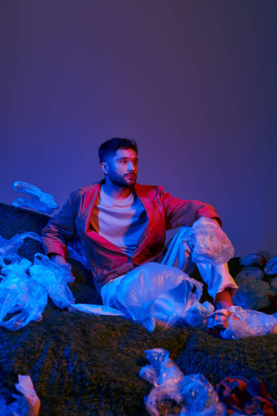 Bir adam, mavi ve kırmızı ışıklarla aydınlatılmış plastik torbalar ve molozların arasında oturur.. - Fotoğraf, Görsel