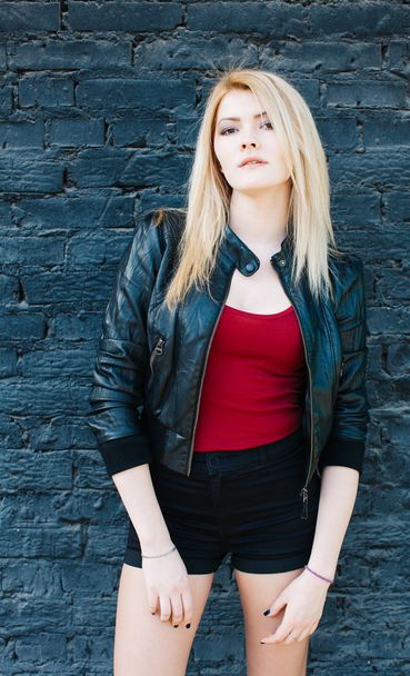 Portrait of a young beautiful blonde girl in a black jacket and shorts posing near brick wall - Φωτογραφία, εικόνα