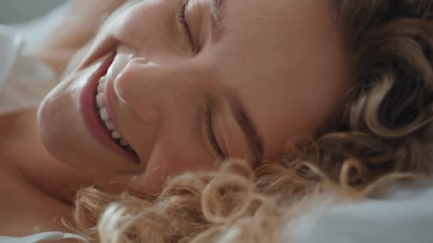 Closeup gentle woman smiling in morning light. Carefree curly lady waking up in bed enjoying peaceful early time. Relaxed beautiful girl rest looking camera at home. Natural beauty femininity concept - Footage, Video