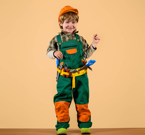 Glimlachende jongen in bouwvakker uniform en veiligheidshelm met gereedschapsriem. gereedschap voor het bouwen. Kleine reparateur met gereedschap voor reparatie. Schattig kind in overall en harde hoed met set van bouwspeelgoed gereedschap - Foto, afbeelding