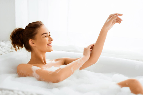 Ευτυχισμένη γυναίκα πλύσιμο του σώματος και να χαλαρώσετε στην μπανιέρα με αφρό - Φωτογραφία, εικόνα
