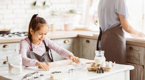 Egy fiatal lány rózsaszín ingben és szürke kötényben tésztát gurít egy fehér pulton a konyhában. Az anyja nem fókuszál a háttérben, más hozzávalókat készít a sütéshez.. - Fotó, kép