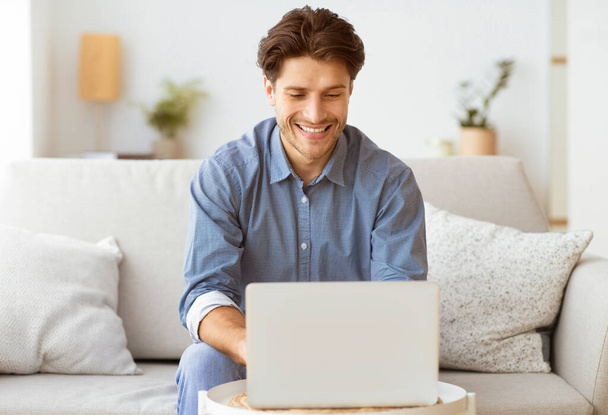 Mężczyzna siedzi na kanapie i uśmiecha się podczas korzystania z laptopa. - Zdjęcie, obraz