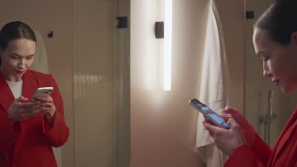 Vonzó nő, aki tükör tükörképet készít. Mosolygó hölgy üzenet a közösségi média csinál szelfi kép okostelefon a fürdőszobában. Stílusos blogger mobiltelefont használ otthon este. - Felvétel, videó