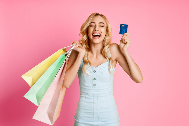Una joven con el pelo largo y rubio sonríe ampliamente mientras sostiene una tarjeta de crédito en una mano y varias bolsas de compras en la otra. Ella está usando un vestido azul claro sobre un fondo rosa. - Foto, imagen