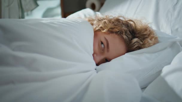 Morning woman resting bedroom in sunlight. Closeup curly girl peeking out blanket enjoying tranquil weekend. Serene blonde looking camera lying stretching on cozy pillow. Smiling model nap at home - Imágenes, Vídeo