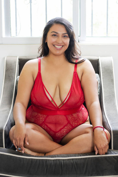 Morena sensual y confiada latina mexicana sentada en sofá vistiendo lencería sonriendo mirando a la cámara - Foto, Imagen