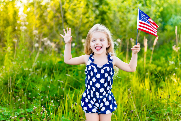 Funny little girl with long curly blond hair putting out her tongue and waving american flag, outdoor portrait on sunny day in summer park. Independence Day, Flag Day concept - Foto, Imagen