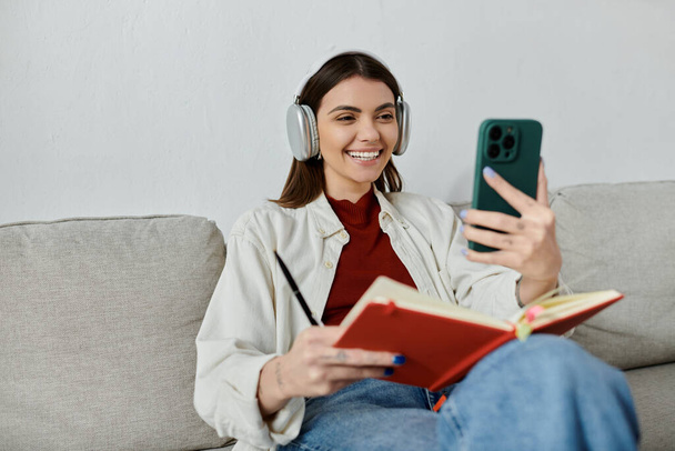 Mladá žena v neformálním oblečení spočívá na pohovce, poslouchá hudbu přes bezdrátová sluchátka, zatímco drží telefon a notebook. - Fotografie, Obrázek