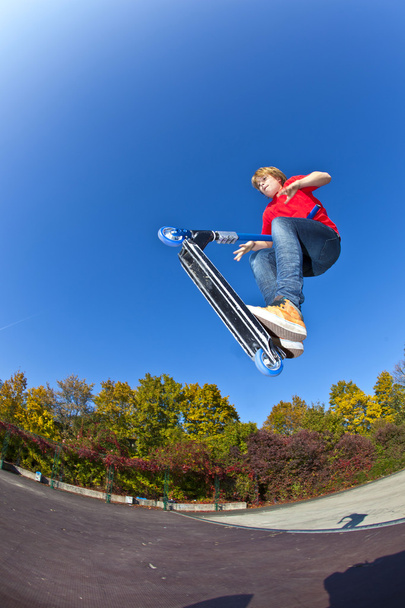 Мальчик прыгает на скутере в скейт-парке под голубым небом.
 - Фото, изображение