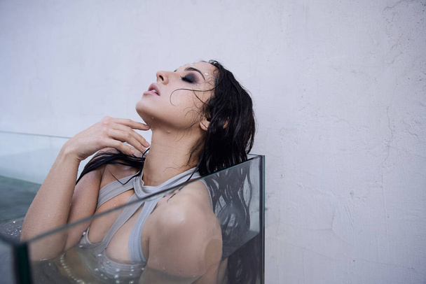 Piękna kobieta z długimi brunetkami unosi się w szklanym zbiorniku wypełnionym wodą, jej oczy zamknięte w kontemplacji. - Zdjęcie, obraz