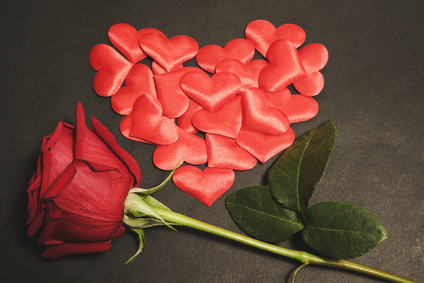 Οριζόντια φωτογραφία από αισθησιακό κόκκινο τριαντάφυλλο και μικρές υφασμάτινες καρδιές, τοποθετημένες σε σχήμα καρδιάς, σε σκούρο μαύρο φόντο. Έννοια Αγίου Βαλεντίνου - Φωτογραφία, εικόνα