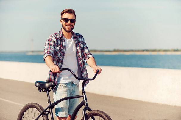 Just me and my bike. Handsome young man standing near his bicycle and smiling while being outdoors - Foto, Bild