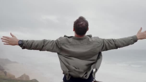Een man strekt zijn armen wijd uit en omarmt het uitgestrekte uitzicht op de oceaan vanaf een klif aan zee op een bewolkte dag, met een gevoel van vrijheid en rust. Man met uitzicht op de oceaan vanaf Cliff Omarmend Vrijheid - Video