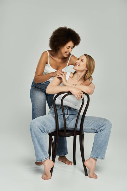 Lesbijska para w białych koszulkach i dżinsach dzieli kochający wzrok siedząc na krześle w studio. - Zdjęcie, obraz