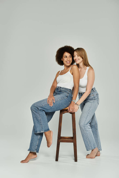 Twee vrouwen, een Black en een White, in jeans en witte topjes glimlachen naar elkaar. Vrouw op kruk heeft voeten op de grond - Foto, afbeelding