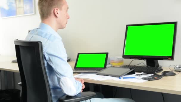 fiatal jóképű férfi ül, és néz az asztali és laptop számítógép az irodában - zöld képernyő - Felvétel, videó