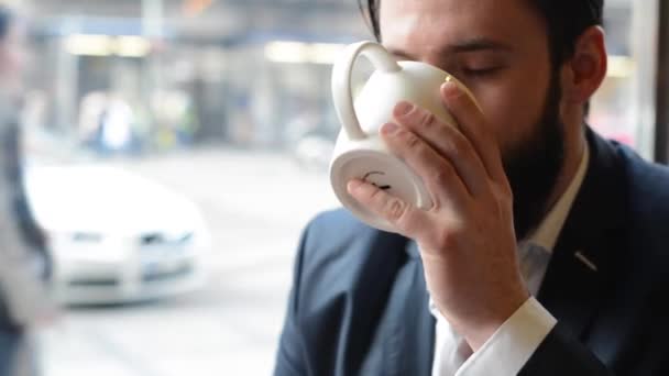 młody przystojny mężczyzna z pełną brodą (hipster) pije kawę w kawiarni - ulica miejska w tle - Materiał filmowy, wideo