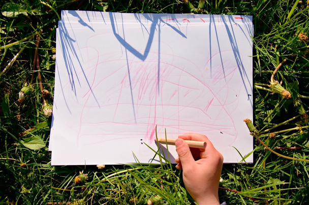 Vue rapprochée de la main de l'enfant dessinant des ornements colorés sur papier. Carnet dans l'herbe verte fraîche de prairie
. - Photo, image