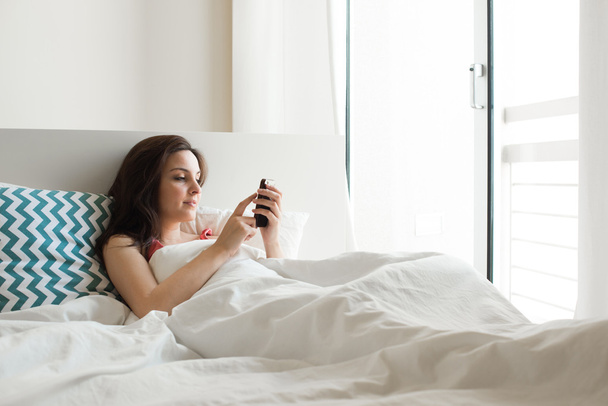 Γυναίκα στο κρεβάτι με smartphone - Φωτογραφία, εικόνα