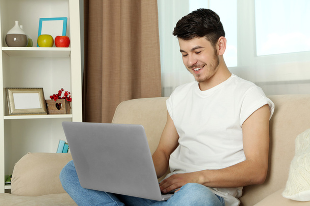 schöner junger Mann sitzt auf Sofa und benutzt Laptop im Zimmer - Foto, Bild