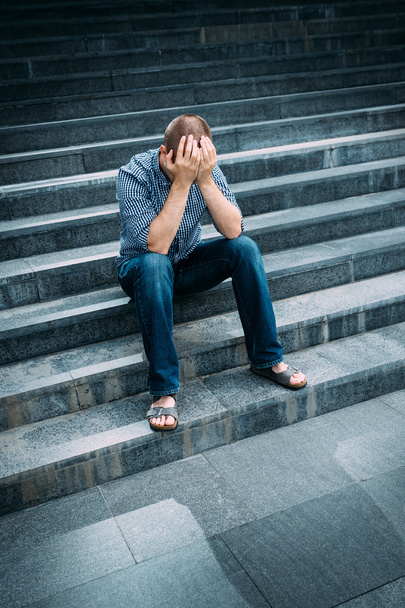 Outdoor portrait of despaired young man covering his face with hands sitting on stairs. Feelings of sadness, despair and tragedy - Foto, afbeelding