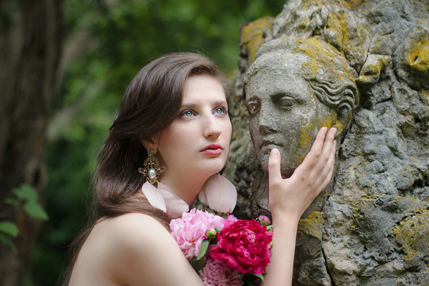 Hermosa mujer desnuda joven con una estatua antigua y ramo de flores. Comparación de la animada belleza femenina y el arte de los antiguos escultores
. - Foto, Imagen