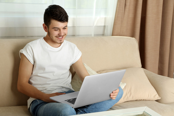 Handsome young man sitting on sofa and using laptop in room - Foto, Bild