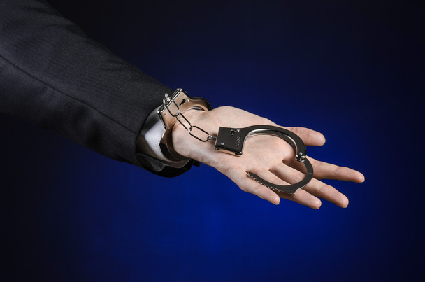 Διαφθορά και δωροδοκία θέμα: επιχειρηματίας σε ένα μαύρο κοστούμι με χειροπέδες στα χέρια του, σε σκούρο μπλε φόντο στο στούντιο απομονωθεί - Φωτογραφία, εικόνα