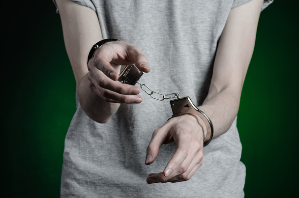 Prison and convicted topic: man with handcuffs on his hands in a gray T-shirt and blue jeans on a dark green background in the studio, put handcuffs on the drug dealer - Foto, imagen
