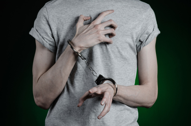 Prison and convicted topic: man with handcuffs on his hands in a gray T-shirt and blue jeans on a dark green background in the studio, put handcuffs on the drug dealer - 写真・画像