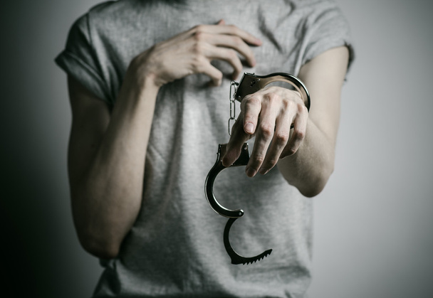 Prison and convicted topic: man with handcuffs on his hands in a gray T-shirt on a gray background in the studio, put handcuffs on rapist - 写真・画像