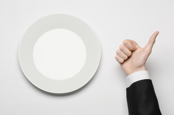 Business lunch and healthy food theme: man's hand in a black suit holding a white empty plate and shows finger gesture on an isolated white background in studio top view - Foto, imagen