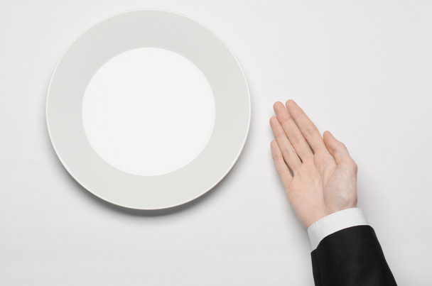 Business lunch and healthy food theme: man's hand in a black suit holding a white empty plate and shows finger gesture on an isolated white background in studio top view - Фото, зображення