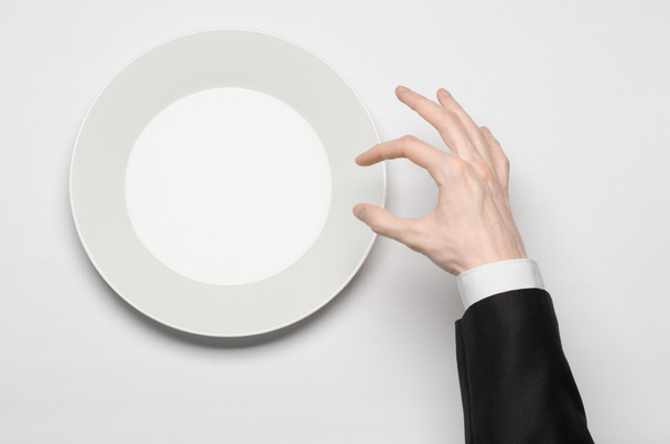 Business lunch and healthy food theme: man's hand in a black suit holding a white empty plate and shows finger gesture on an isolated white background in studio top view - Photo, Image