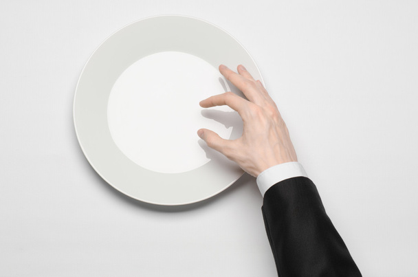 Business lunch and healthy food theme: man's hand in a black suit holding a white empty plate and shows finger gesture on an isolated white background in studio top view - Foto, Bild