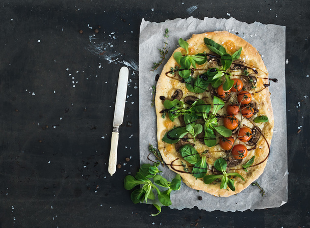 Rustic homemade pizza with fresh lambs lettuce, mushrooms and cherry-tomatoes over dark grunge background - Photo, image