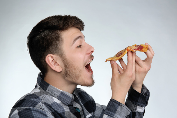Young man eating piece of pizza on gray background - Fotó, kép