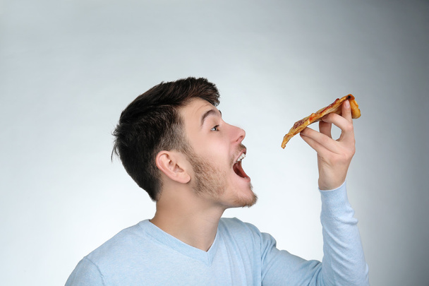 Young man eating piece of pizza on gray background - Zdjęcie, obraz