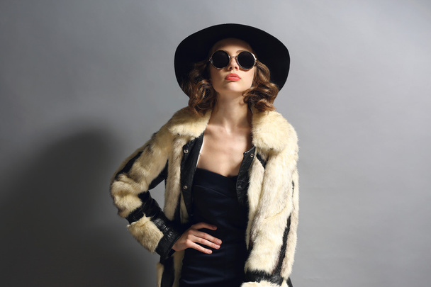 Portrait of beautiful model in fur coat, hat and sunglasses on gray background - Photo, Image