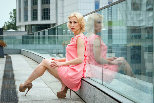 Outdoor summer fashion stunning portrait on pretty young blonde sexy woman dressed in pink dress wearing pastel colored jewelry on the background of the urban landscape. - Photo, Image