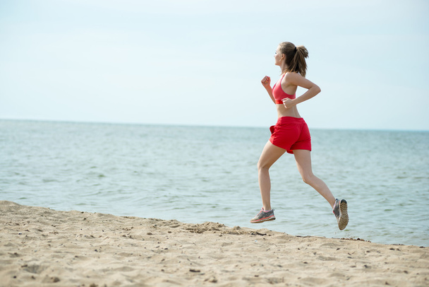 Νεαρή κοπέλα που τρέχει στην παραλία άμμου ηλιόλουστο καλοκαίρι. Προπόνηση. Σπρώχνω - Φωτογραφία, εικόνα