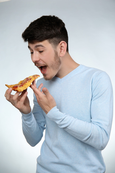 Young man eating piece of pizza on gray background - Fotoğraf, Görsel