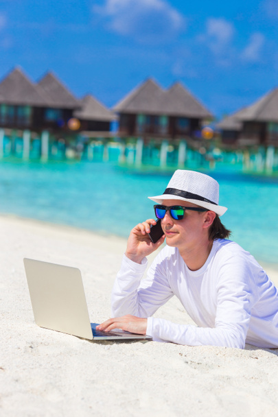 Nuori liikemies tietokoneen ja matkapuhelimen kanssa valkoisella rannalla
 - Valokuva, kuva